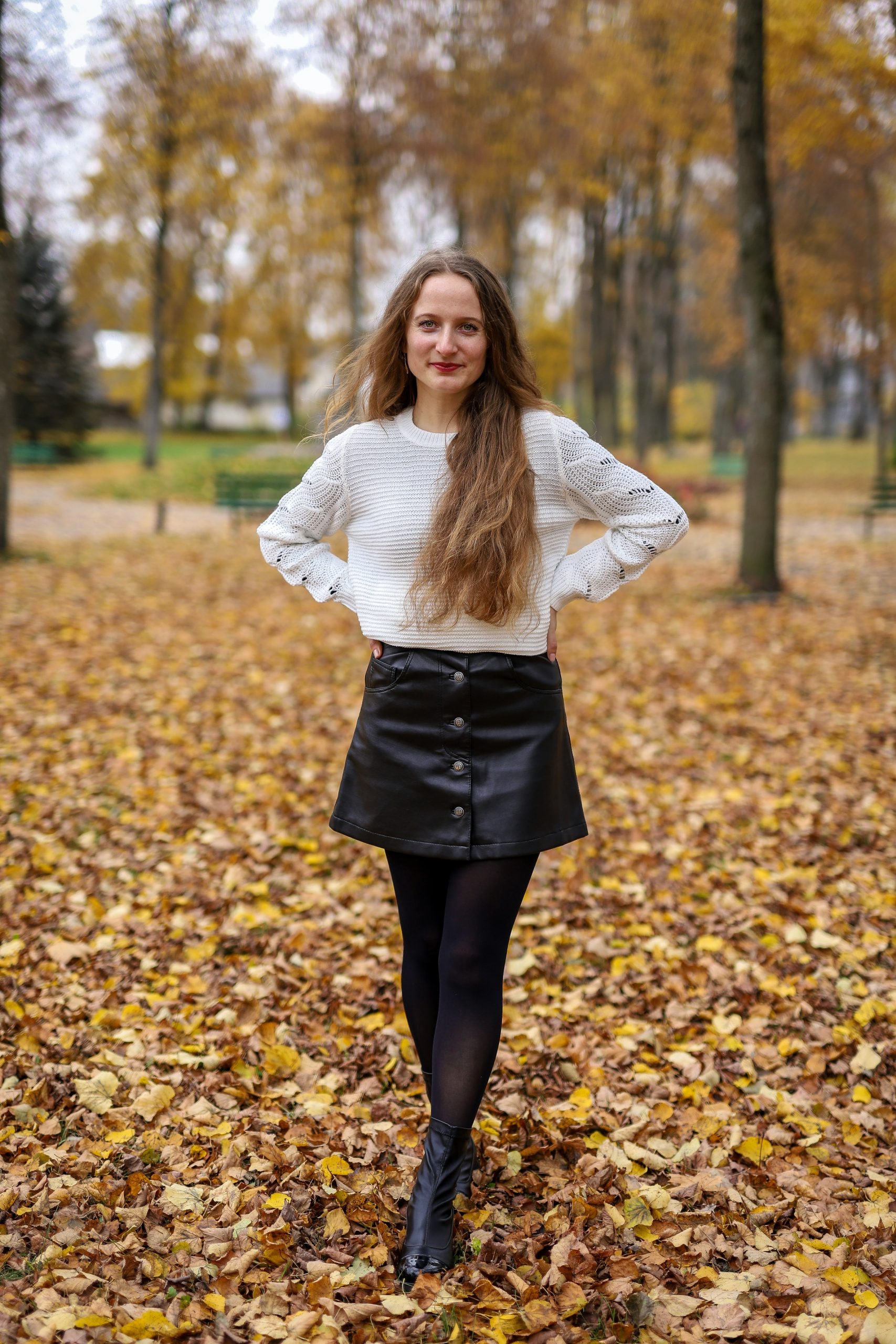 A woman with a confident smile stands in a pile of golden autumn leaves. She wears a skirt and has one hand on her waist. The leaves surround her, creating a warm and inviting atmosphere.